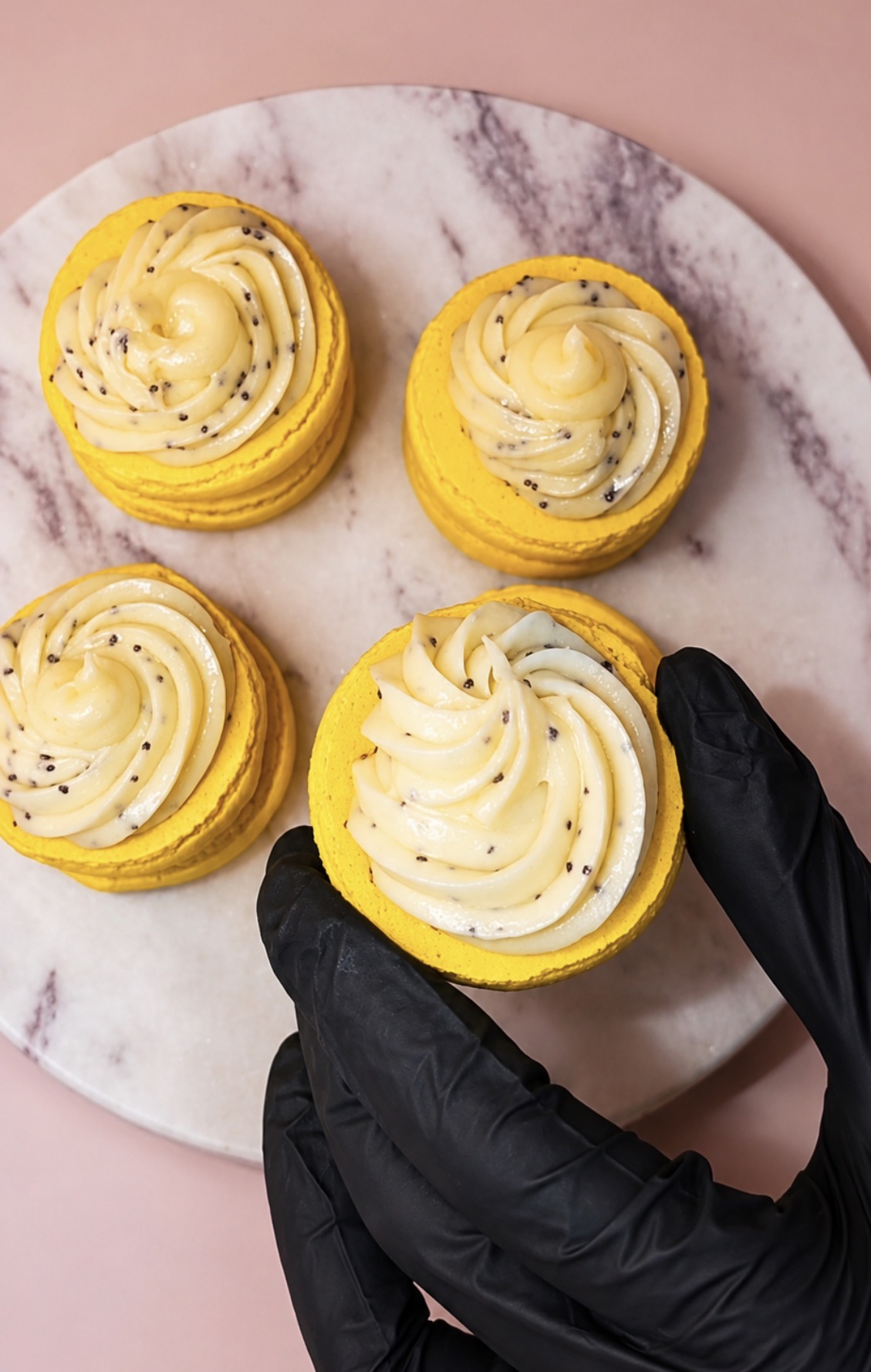 Macarons citron graine de pavot tenus par une main gantée au-dessus d'un plateau en marbre